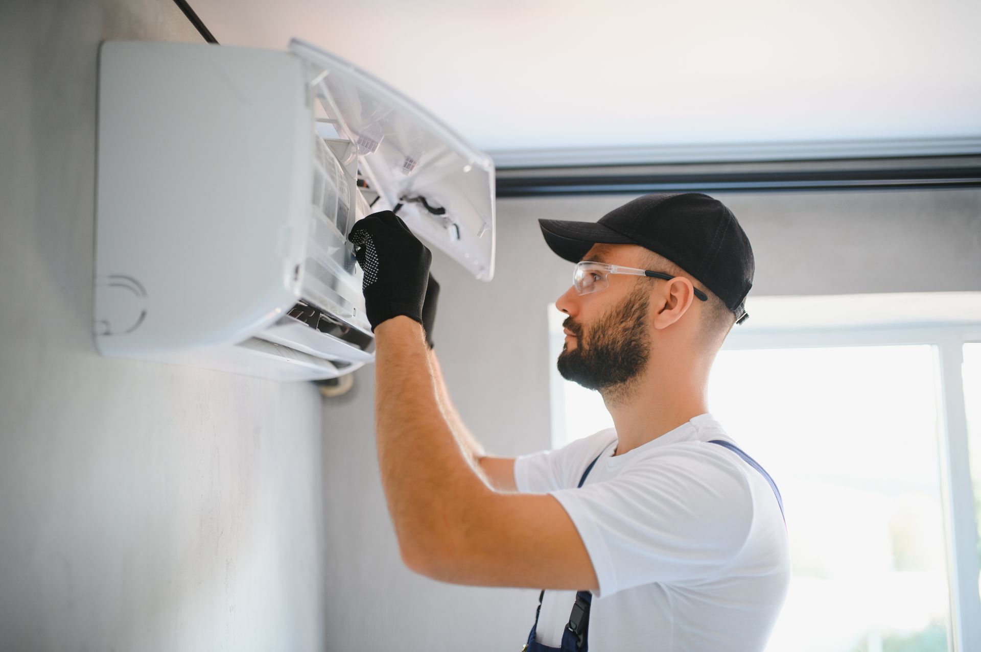 Man in a baseball cap and safety glasses servicing a wall-mounted air conditioning unit.