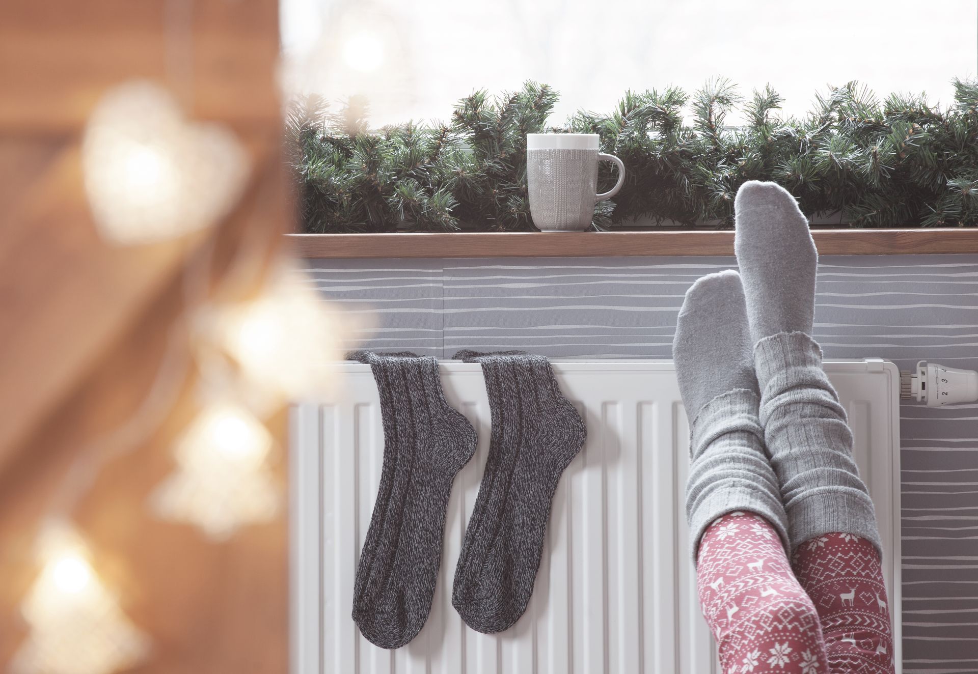 Person with cozy socks up on a radiator, with a cup, window with garland, and Christmas lights.
