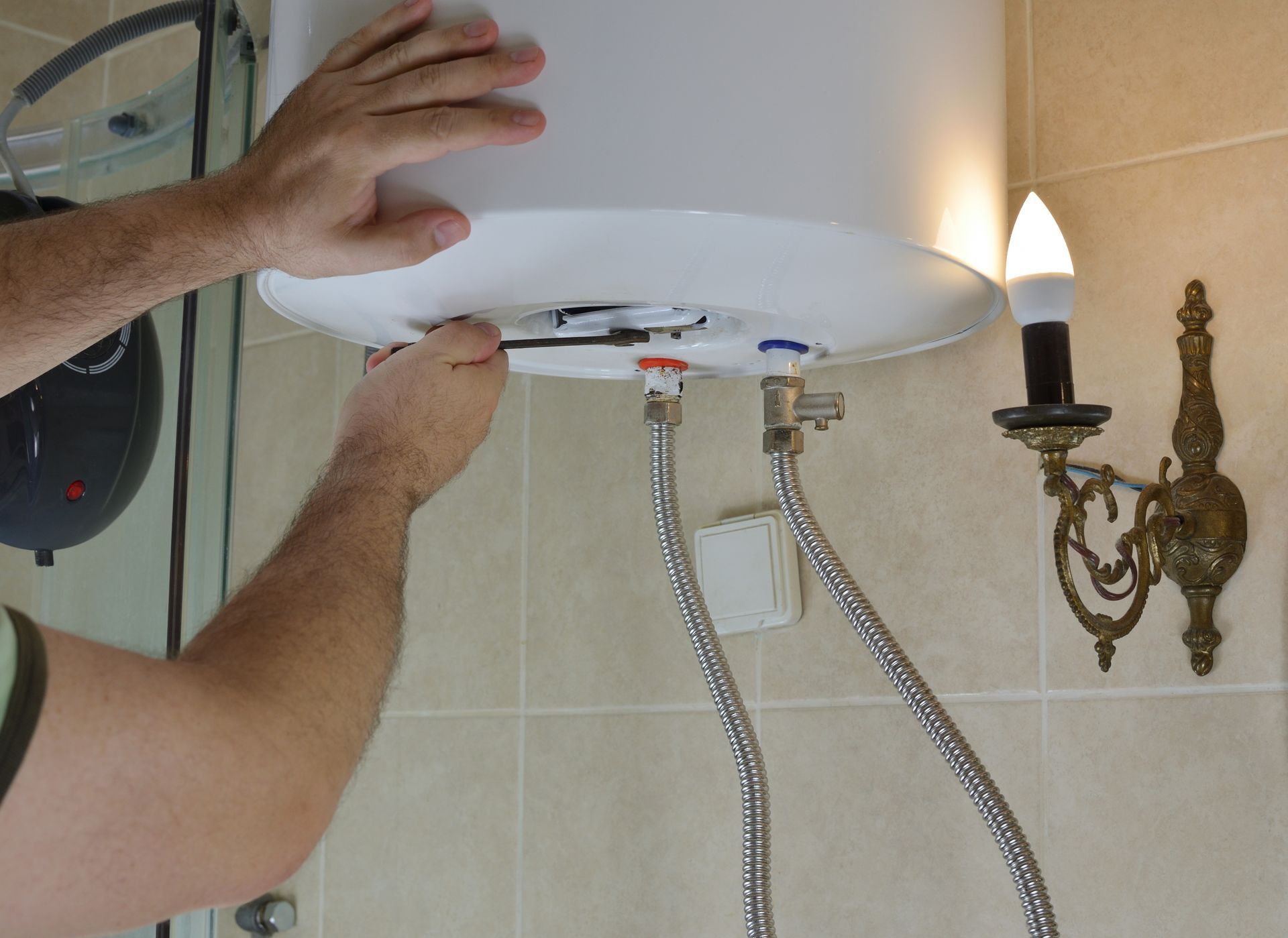 Hands using a wrench to repair a white wall-mounted water heater in a tiled bathroom.