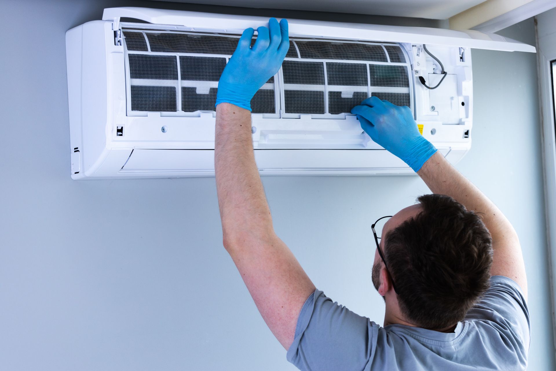 Man in blue gloves cleaning an air conditioner filter mounted on a light blue wall.