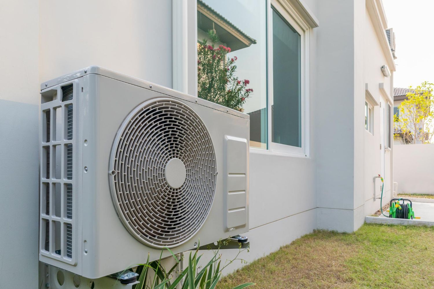 A white air conditioner is mounted on the side of a house next to a window.