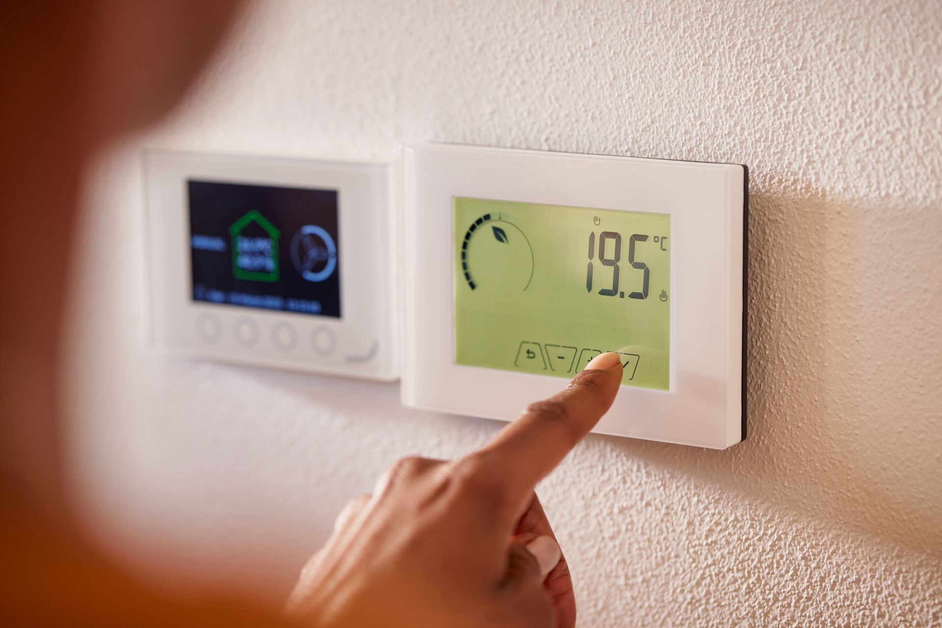 A person's hand adjusting a digital thermostat on a white wall, set to 19.5 degrees Celsius.