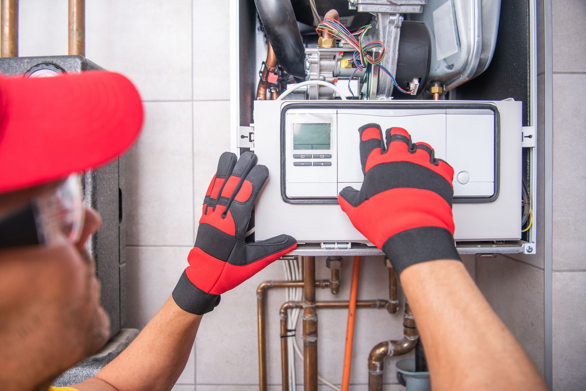 Person in red cap and gloves servicing a furnace, indoors.
