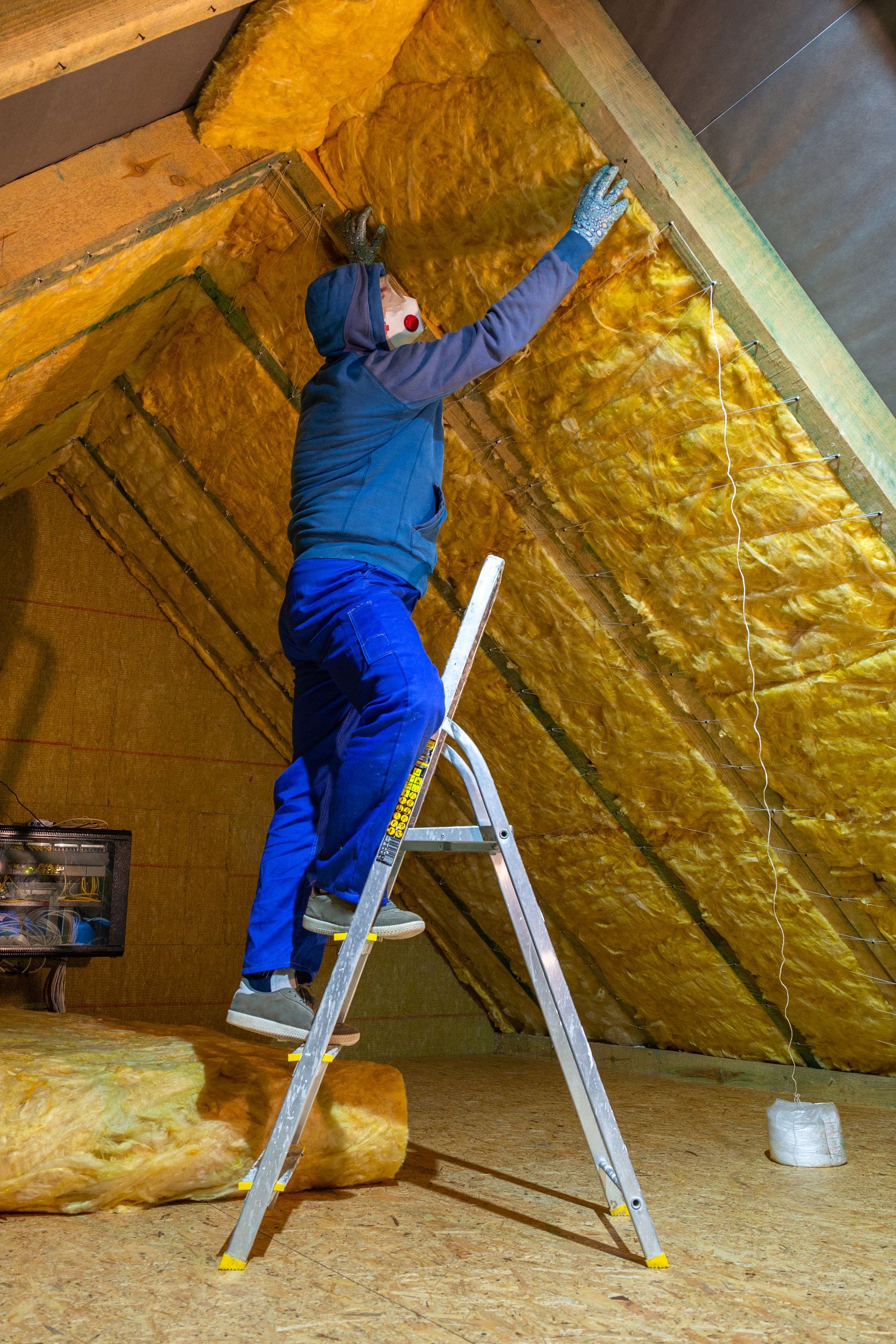 A man is standing on a ladder in an attic insulating the ceiling.