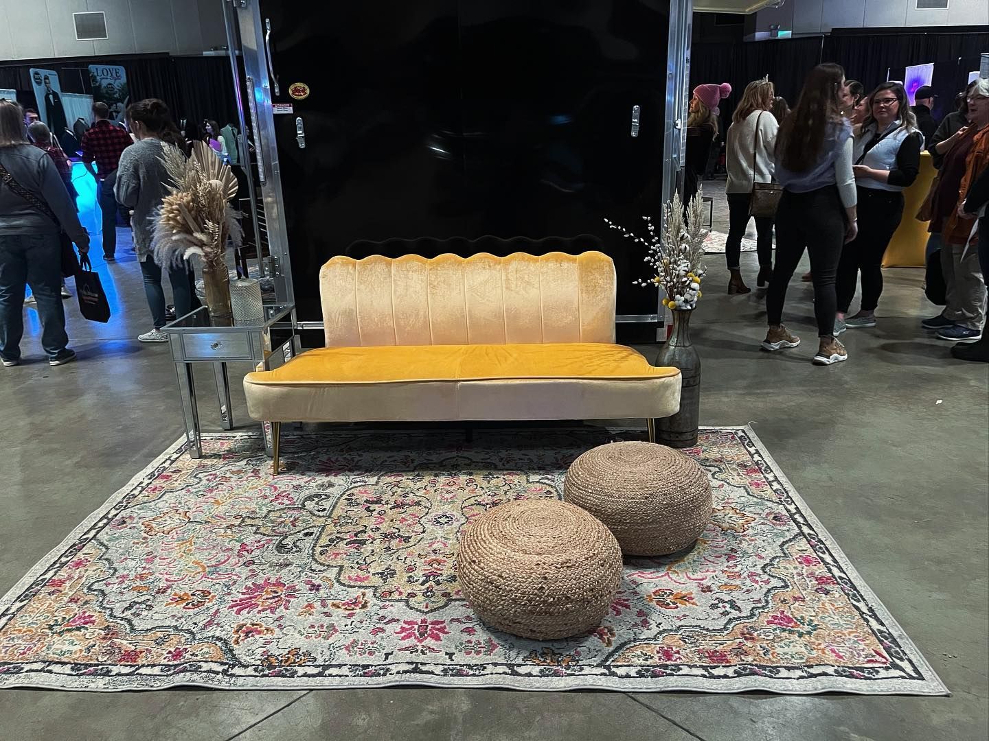 A yellow velvet sofa on a rug, with two round woven ottomans in front, inside an event. People in the background.