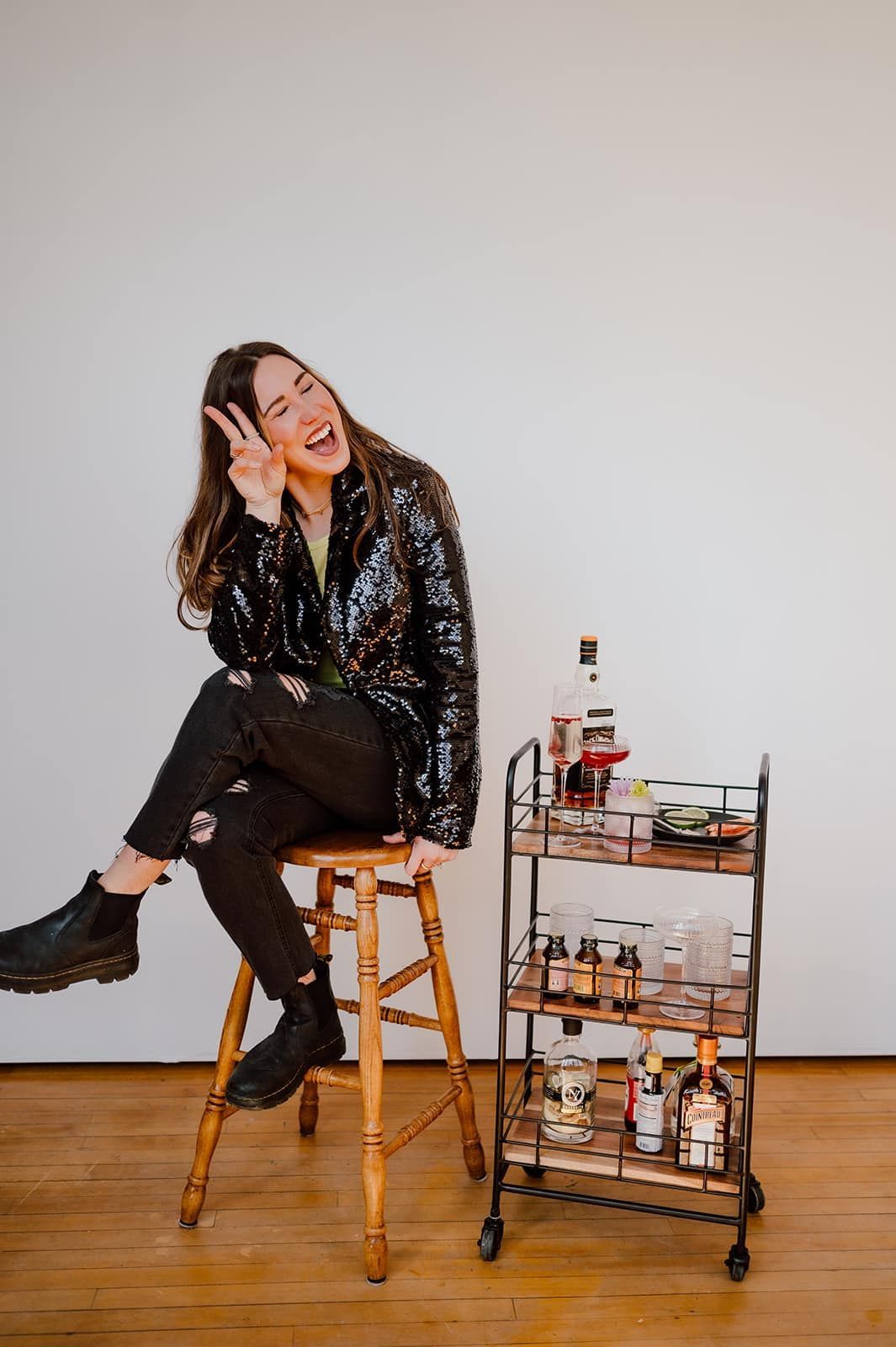 Woman seated, smiling, making peace sign; next to a bar cart with bottles and glasses.