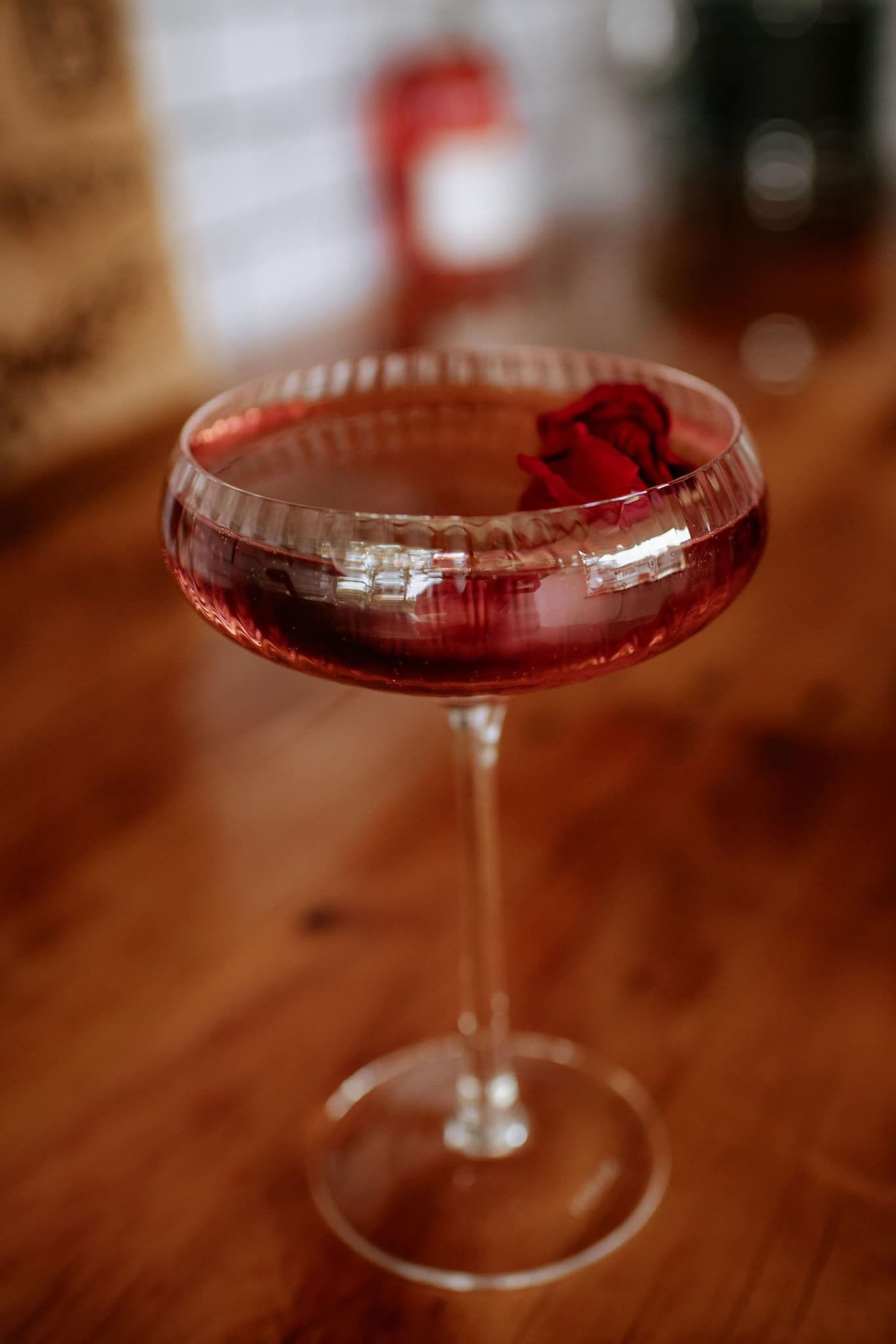 Cocktail in a coupe glass, garnished with a red flower, on a wooden surface.