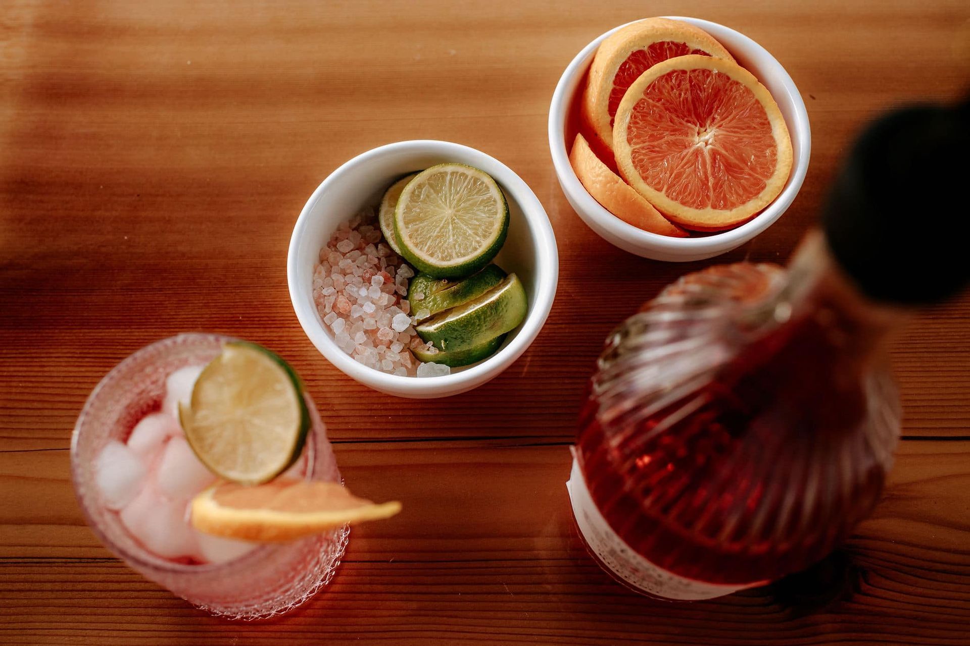 Cocktail ingredients: sliced oranges, limes, pink salt, with a drink and a bottle on wood.