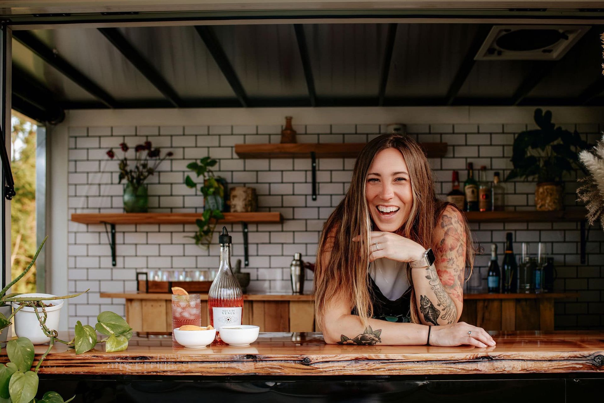 Woman smiling behind a bar with brick backsplash. Wooden shelves, plants, and bottles are visible.