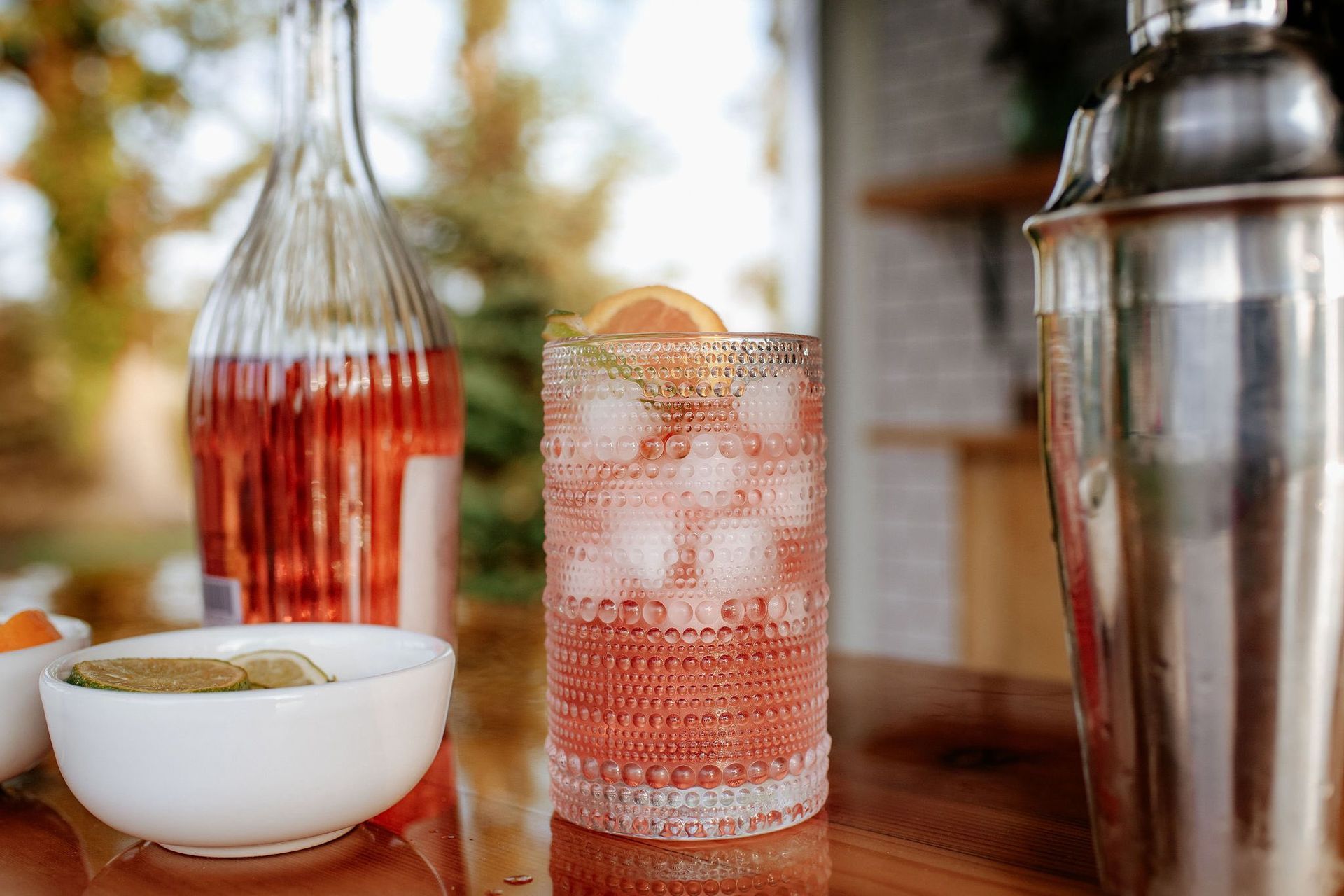 Cocktail with ice, grapefruit slice, pink liquid, beside a bottle and shaker on a wooden surface.