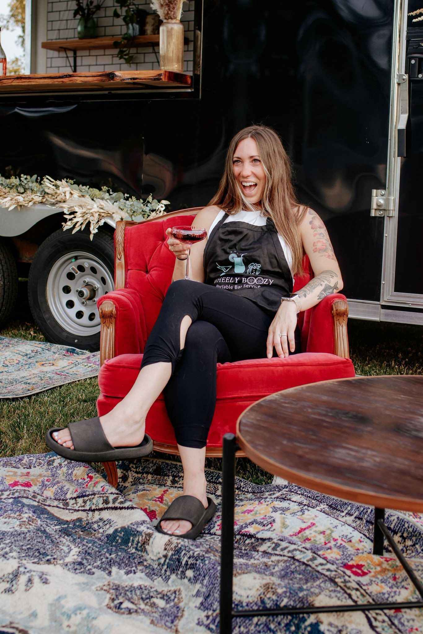 Woman in apron, holding wine, sitting in red chair outdoors, smiling near a trailer.