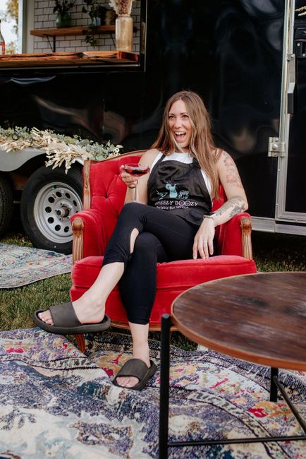 Woman in apron, holding wine, sitting in red chair outdoors, smiling near a trailer.