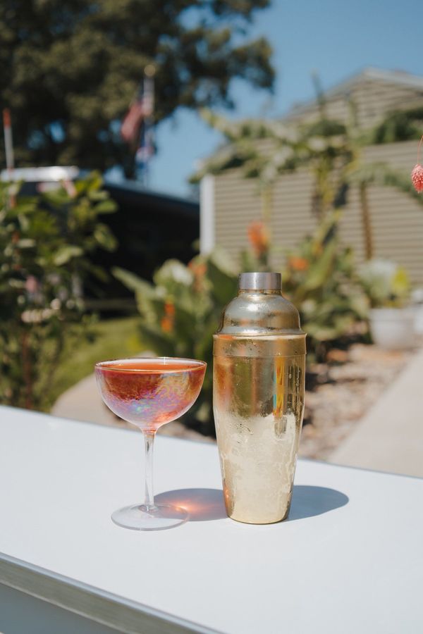 Golden cocktail shaker and iridescent glass on a white surface, outdoor setting with greenery.