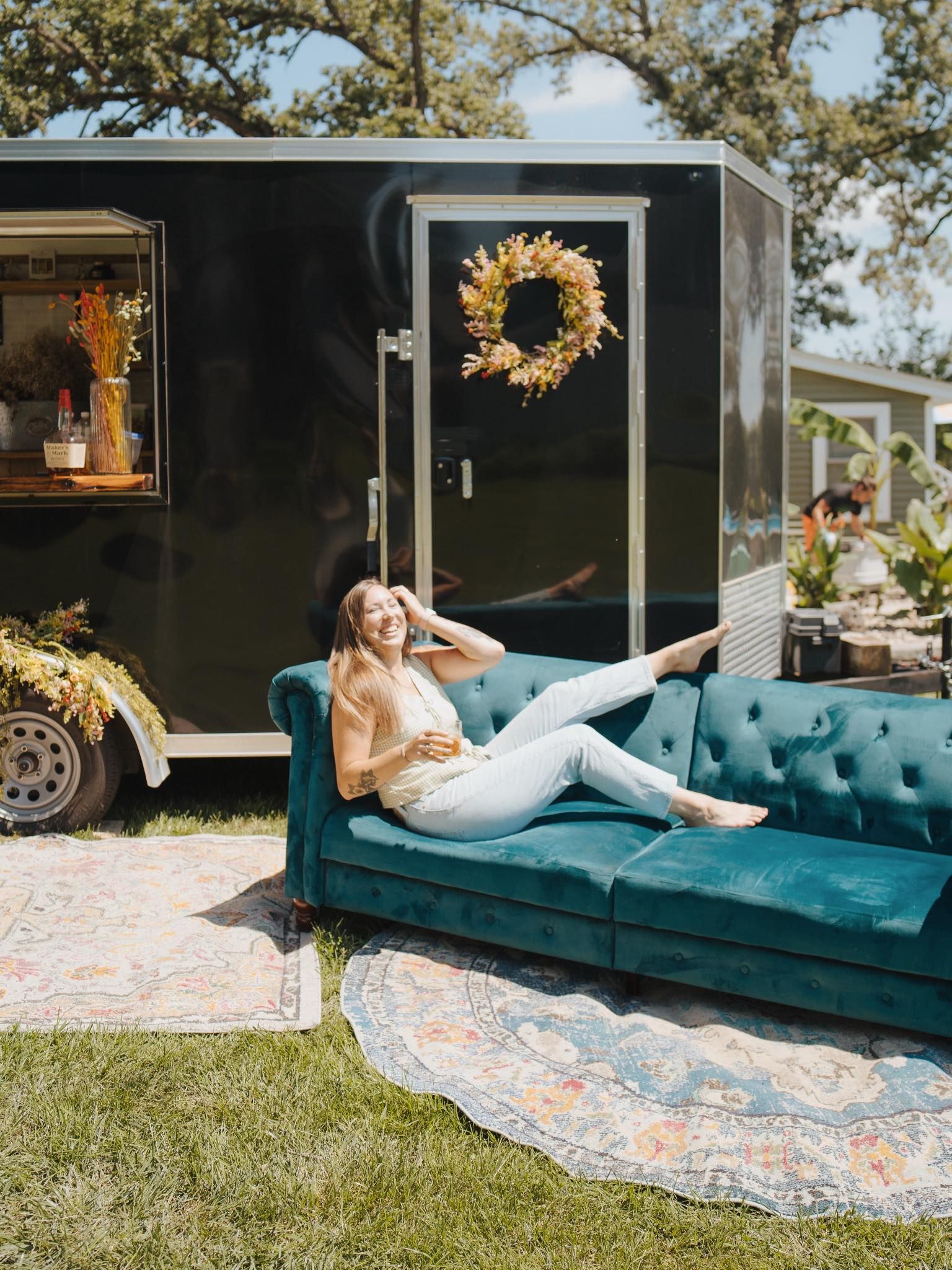Woman reclines on teal couch outside black trailer, smiling. A wreath decorates the door.