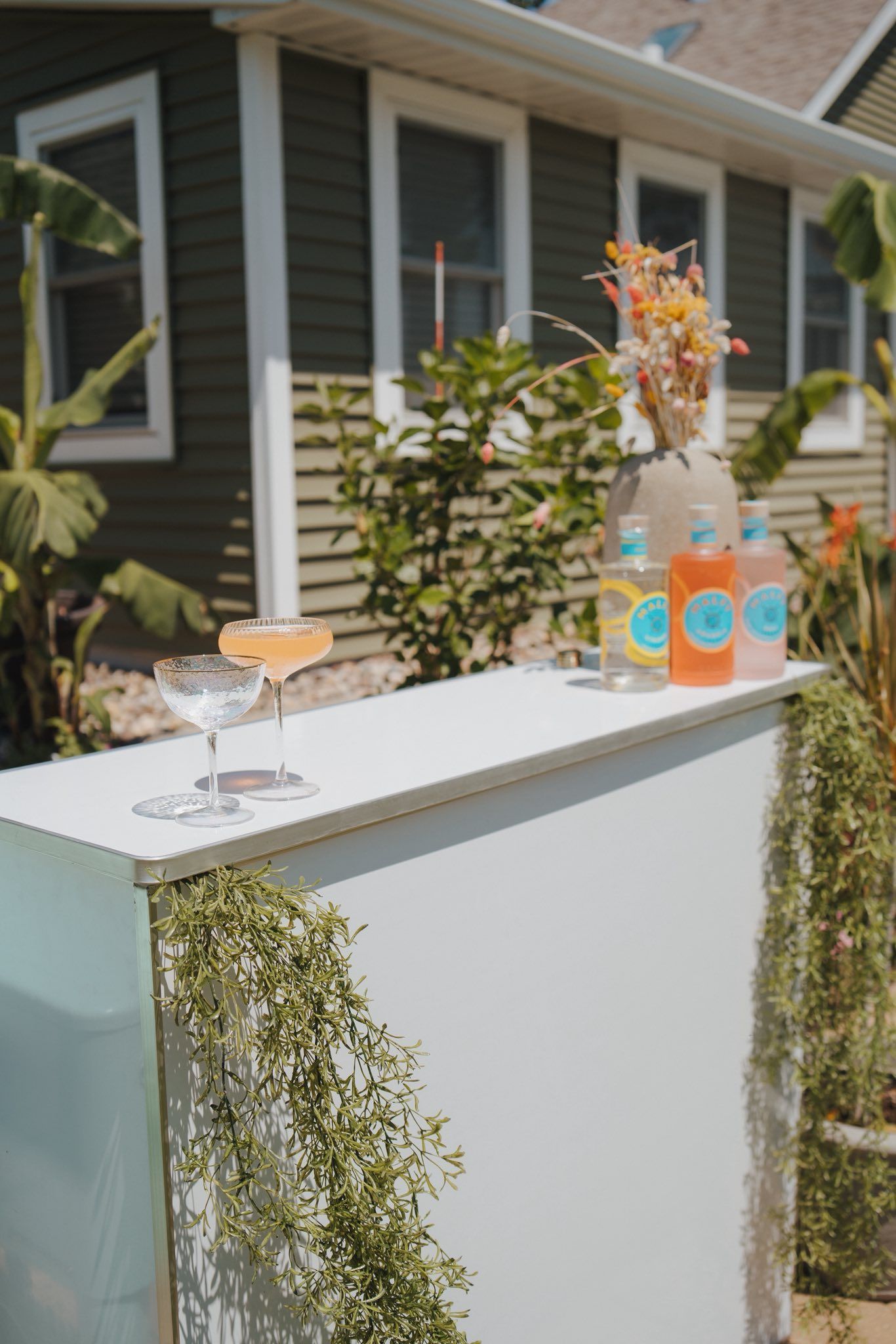 Outdoor bar with cocktails and bottles, decorated with greenery and flowers.