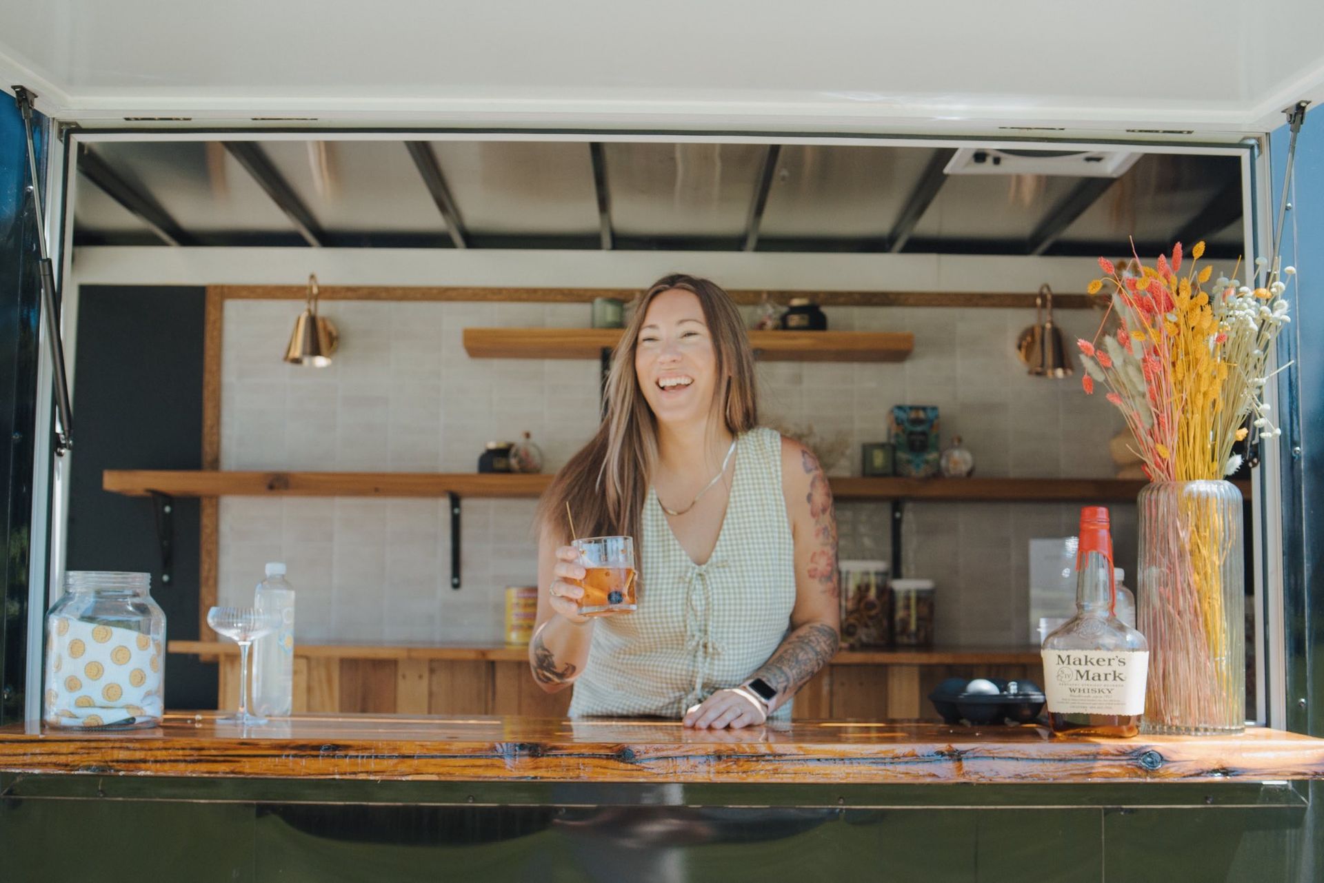 Woman behind a mobile bar, holding a drink, smiling. Wooden bar, gold accents, colorful flowers.