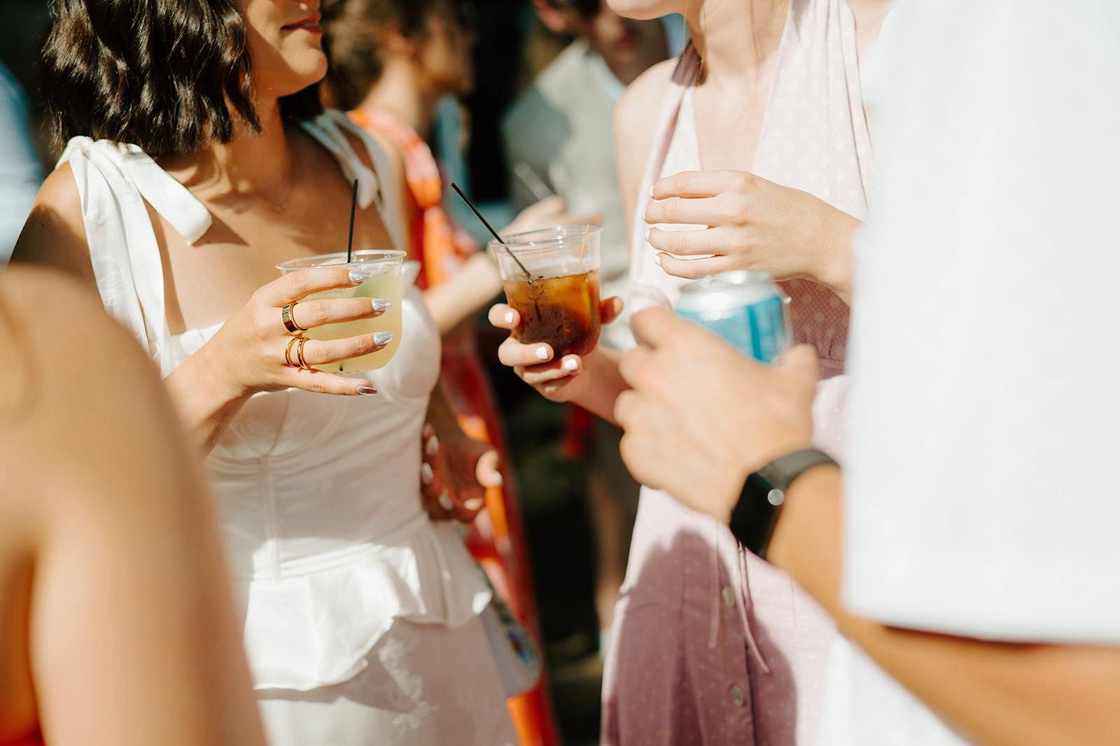 People at an outdoor gathering, holding drinks, light colors, sunlight.