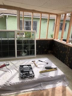 Room under construction, with windows and tools on a sheet-covered surface, wooden frame, and green houses in background.