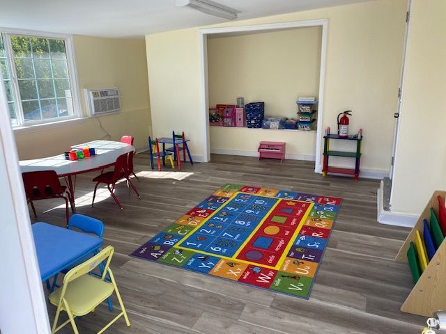 A brightly lit playroom with tables, chairs, a rug, toys, and a storage nook.