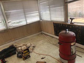 A red smoker in a sunroom under renovation; dirty floor, damaged wall, blinds closed.