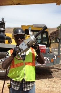 Construction worker in safety vest and hard hat holding a large wrench, smiling outdoors.