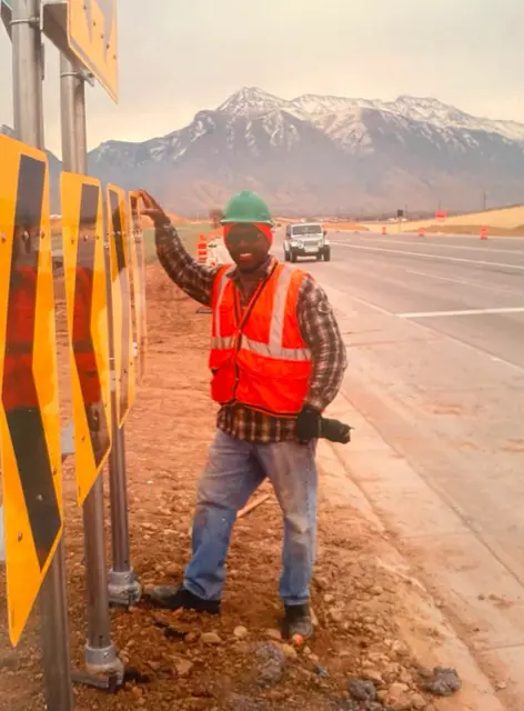 Construction worker by road signs, mountain backdrop. Man in safety vest smiles, leaning, holding tool.