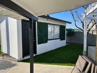 White shed with black door and green accent, next to a patio and artificial turf.