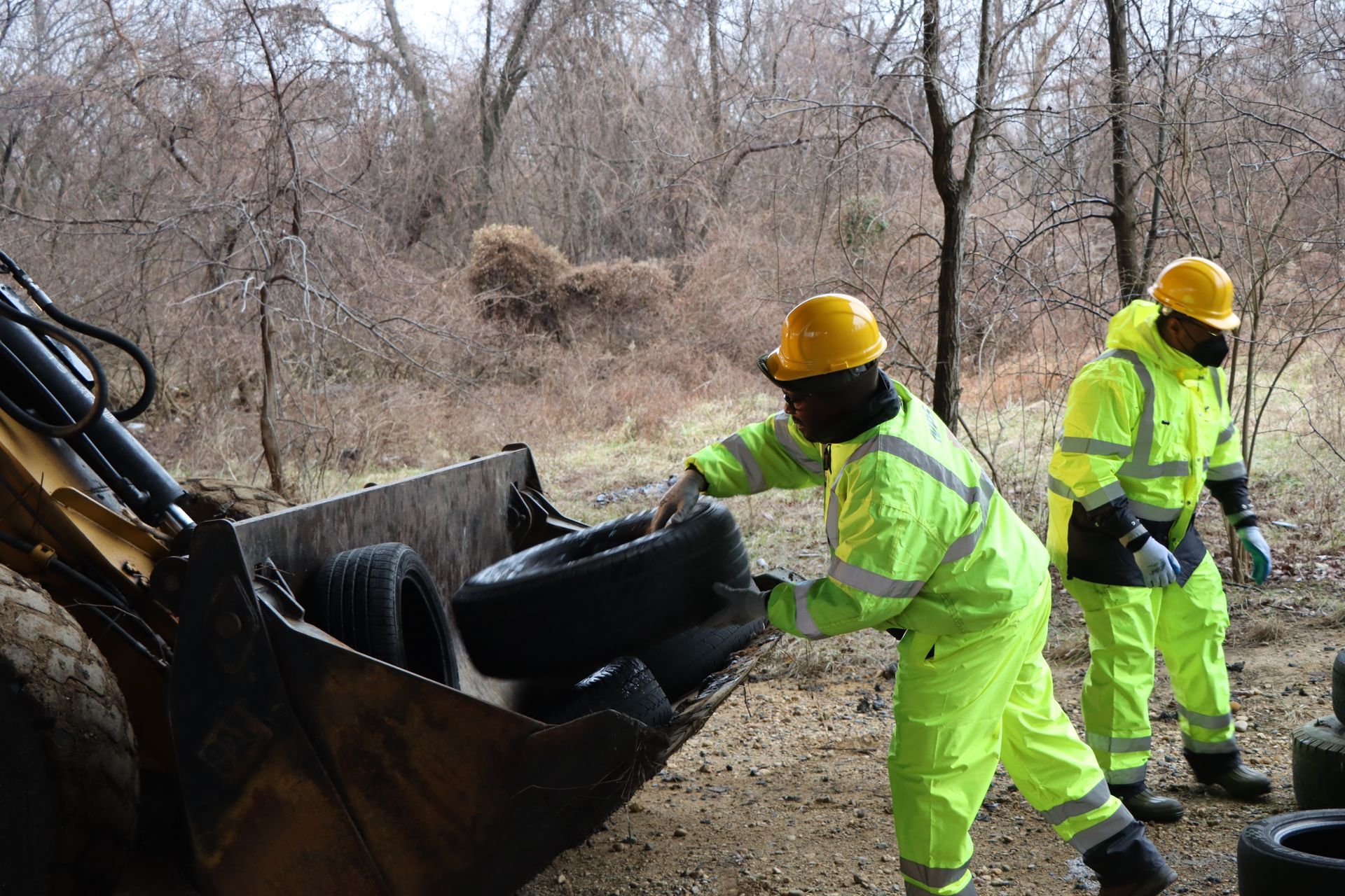 Two men in yellow safety suits are loading tires into a bucket.