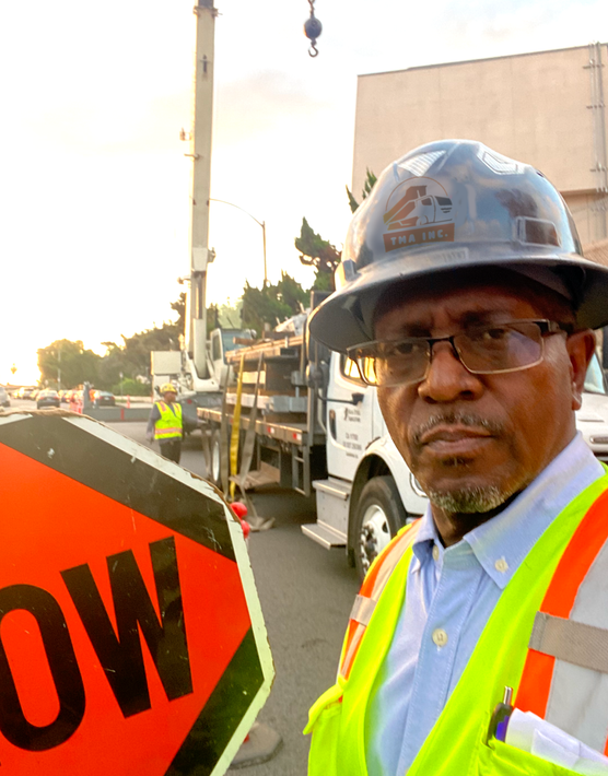 A man wearing a hard hat stands next to an orange sign that says slow