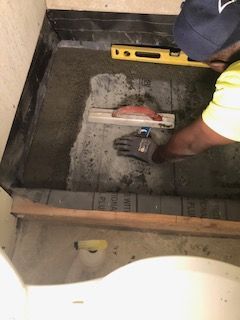 Person leveling concrete in a shower base, using a level. Yellow shirt, gray gloves, and a blue hat.