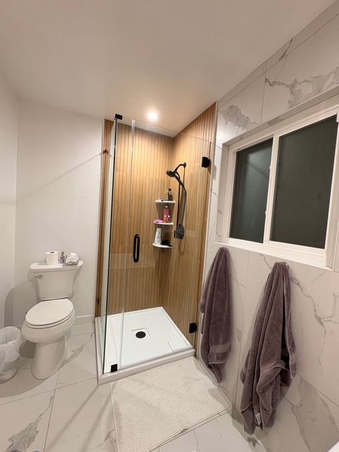 Modern bathroom with a glass shower, toilet, and marble-look tile. Two towels hang on the wall near the window.