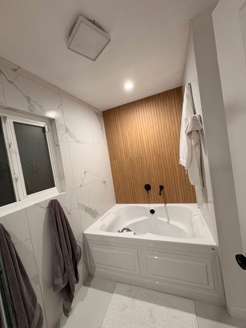 A modern bathroom with a white tub, marble walls, and a wood accent wall; towels hang nearby.