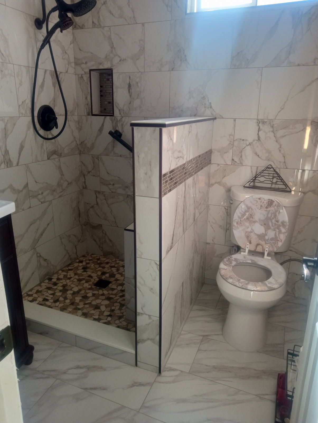 Bathroom with marble-look tiles, shower, toilet with decorative seat, and a partial divider wall.