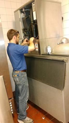 Man in blue shirt repairs ice machine in a commercial kitchen setting.