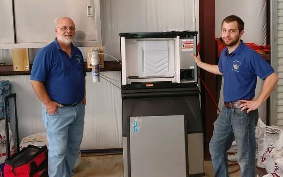 Two men in blue shirts stand next to a commercial ice machine. They are indoors.