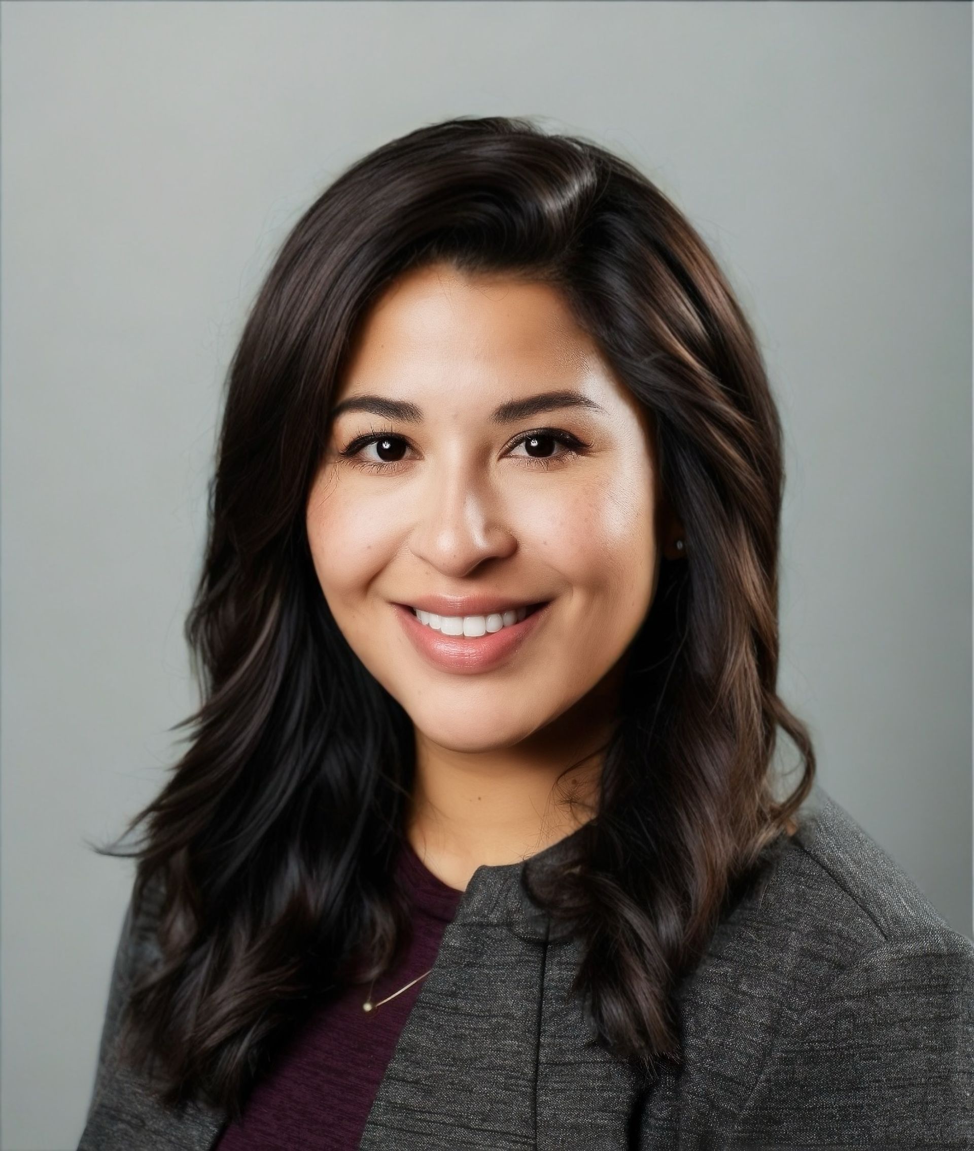 A woman with long dark hair is smiling and wearing a grey jacket