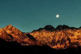 A full moon is rising over a snowy mountain range.