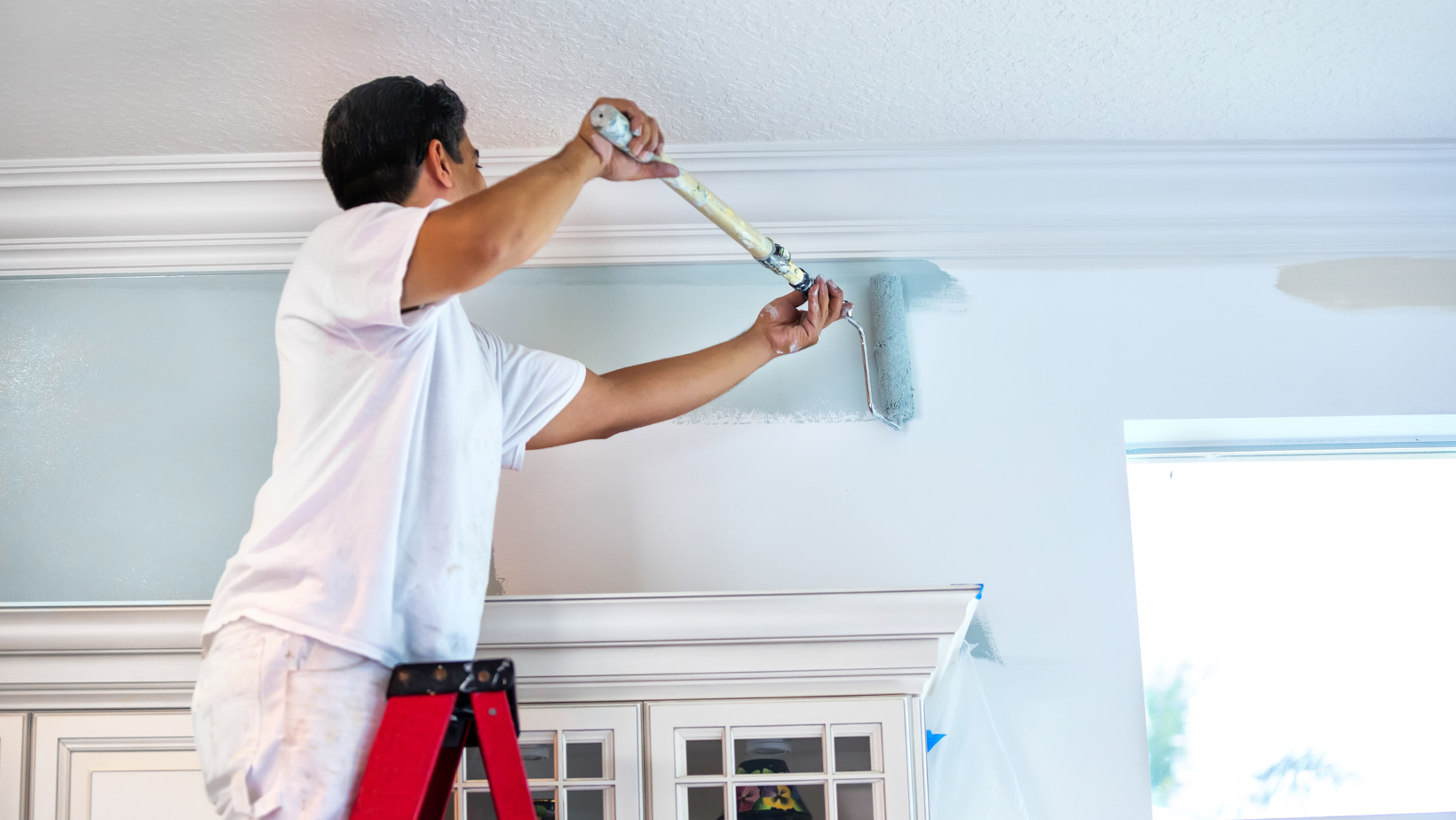 Man paints a light blue wall with a roller while standing on a red stepladder inside a home.