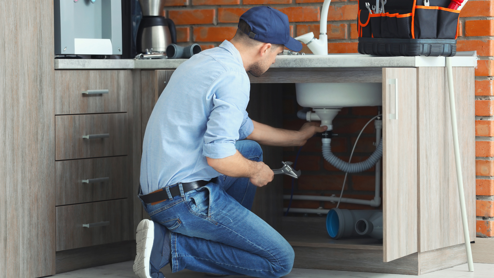 Plumber kneeling under a kitchen sink, working on the pipes. Tools, brick wall background.