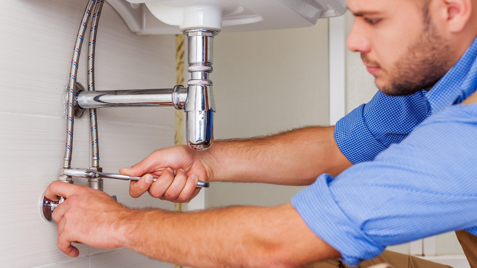Plumber tightening a faucet connection under a white sink with a wrench. Blue shirt, working indoors.