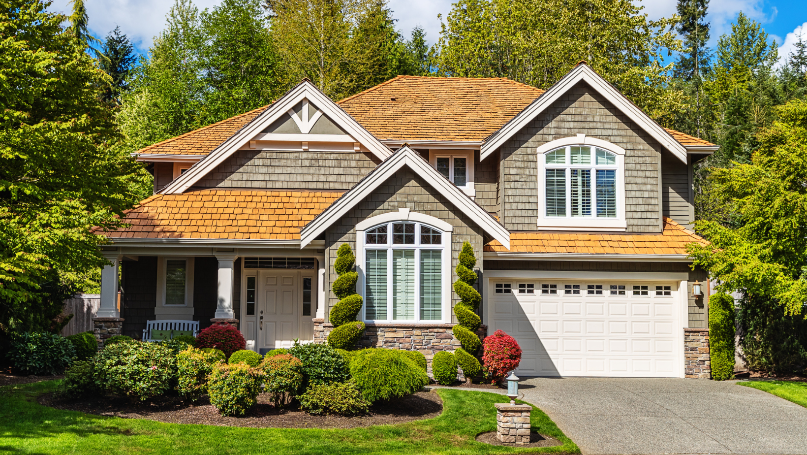 Two-story house with tan roof and gray siding surrounded by green trees and shrubs.