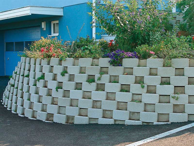 Eine Backsteinmauer mit Blumen darauf vor einem blauen Gebäude