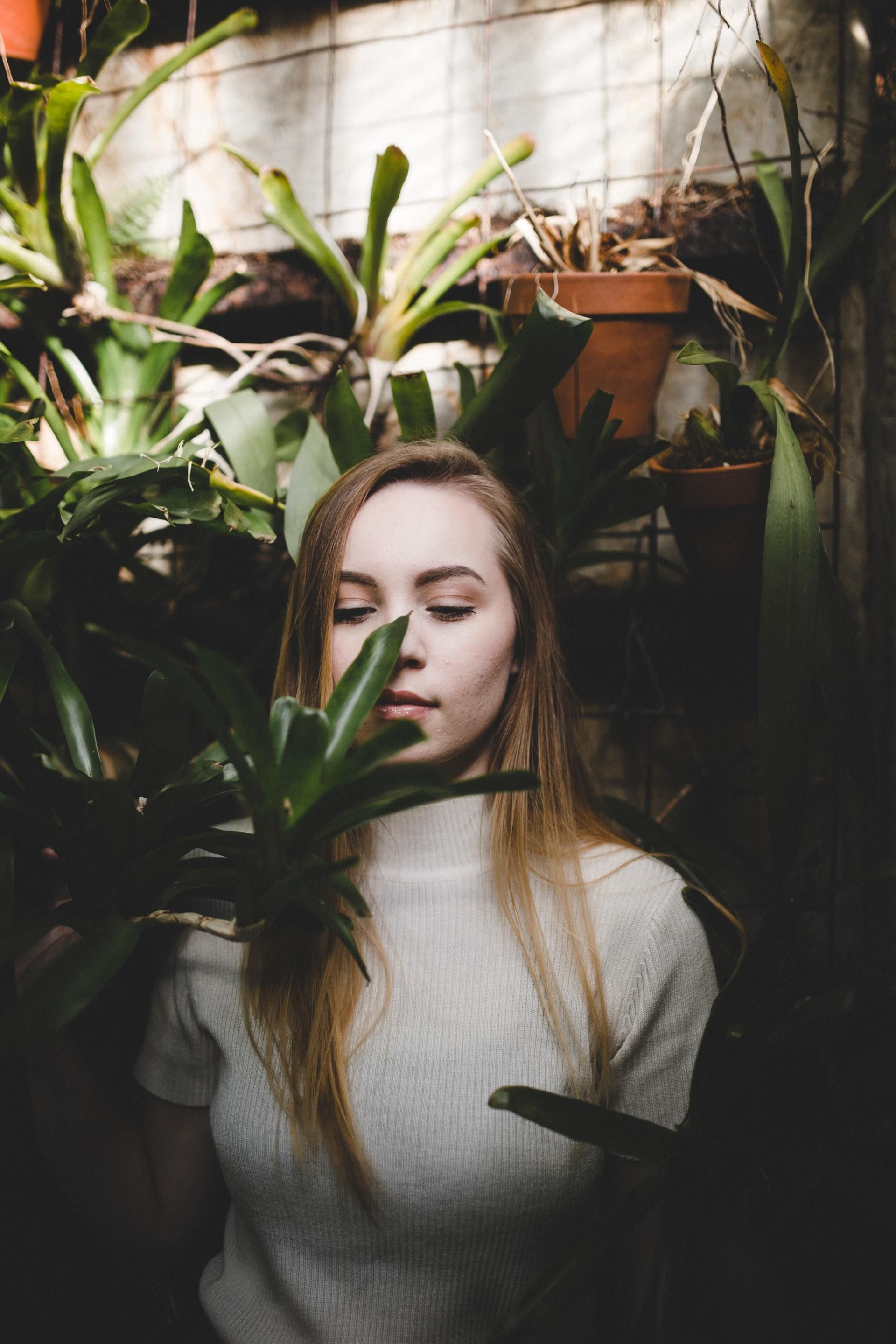 A person stands amidst lush green potted plants, holding a leaf near their face with a serene expression.
