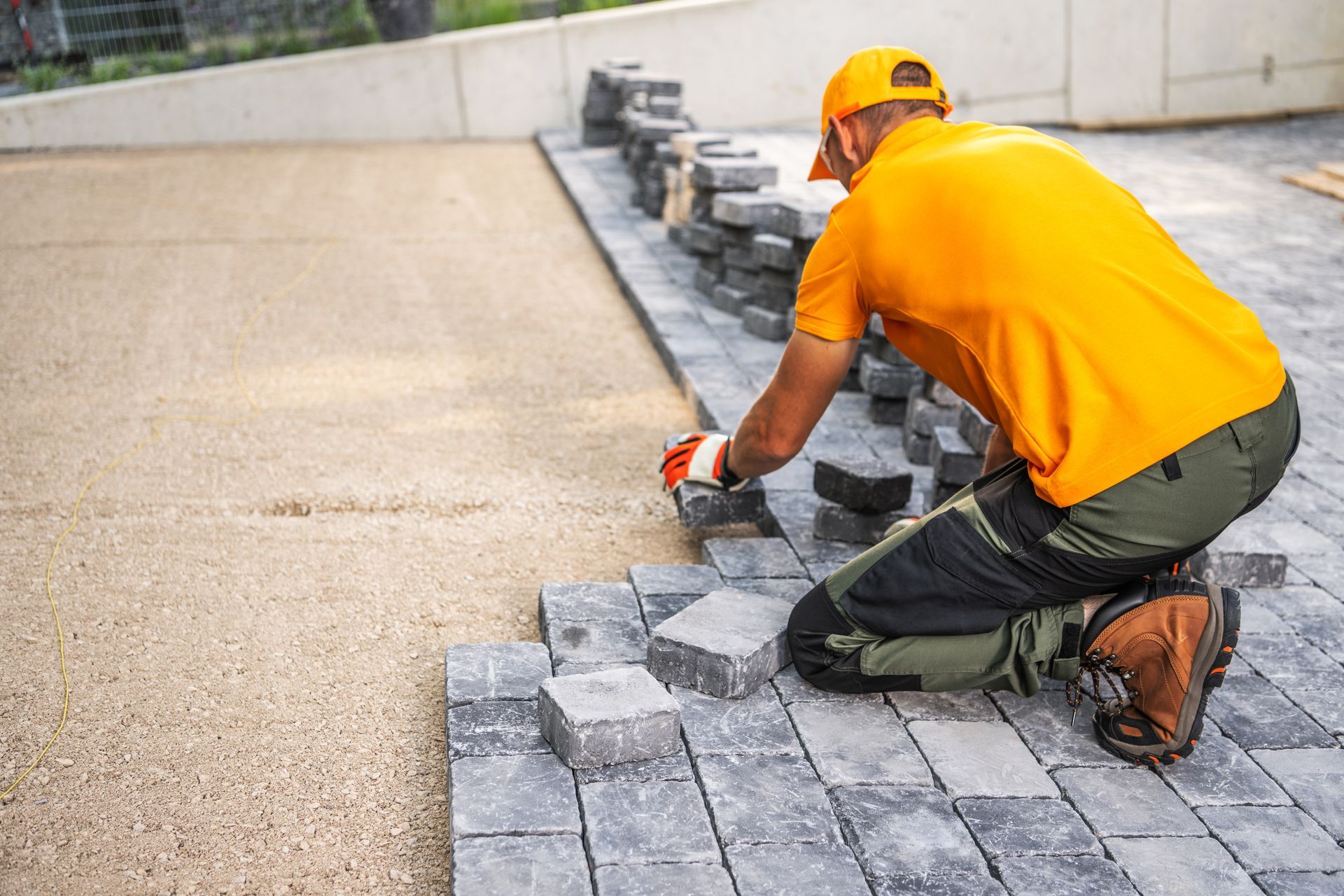 A worker in a yellow shirt and work pants kneels, placing grey paving stones onto a prepared sandy surface.