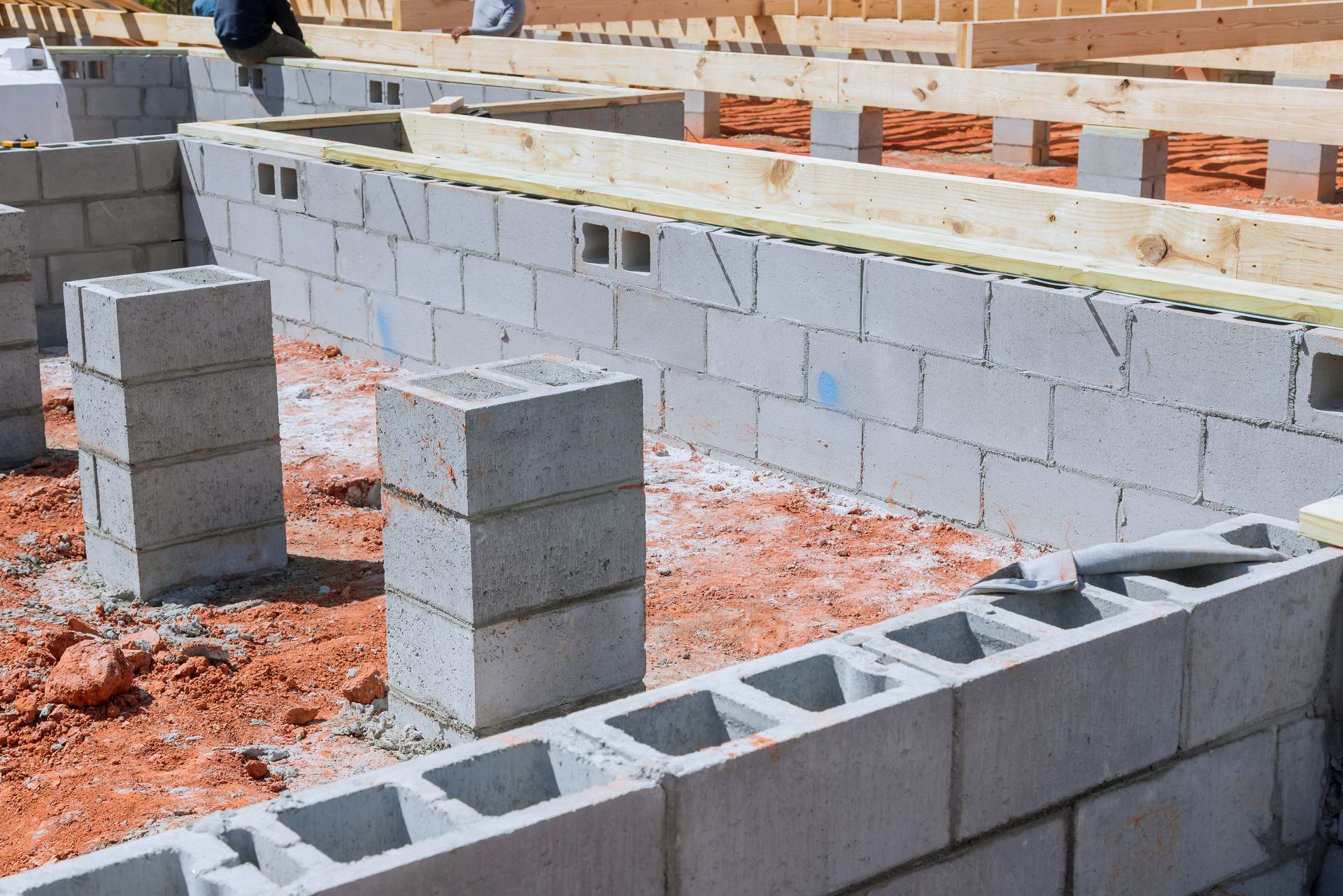 A construction site showing concrete block foundation walls and support piers being built on red dirt soil.