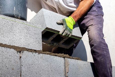 A construction worker in a yellow glove lays a gray concrete block onto a mortar-covered wall.