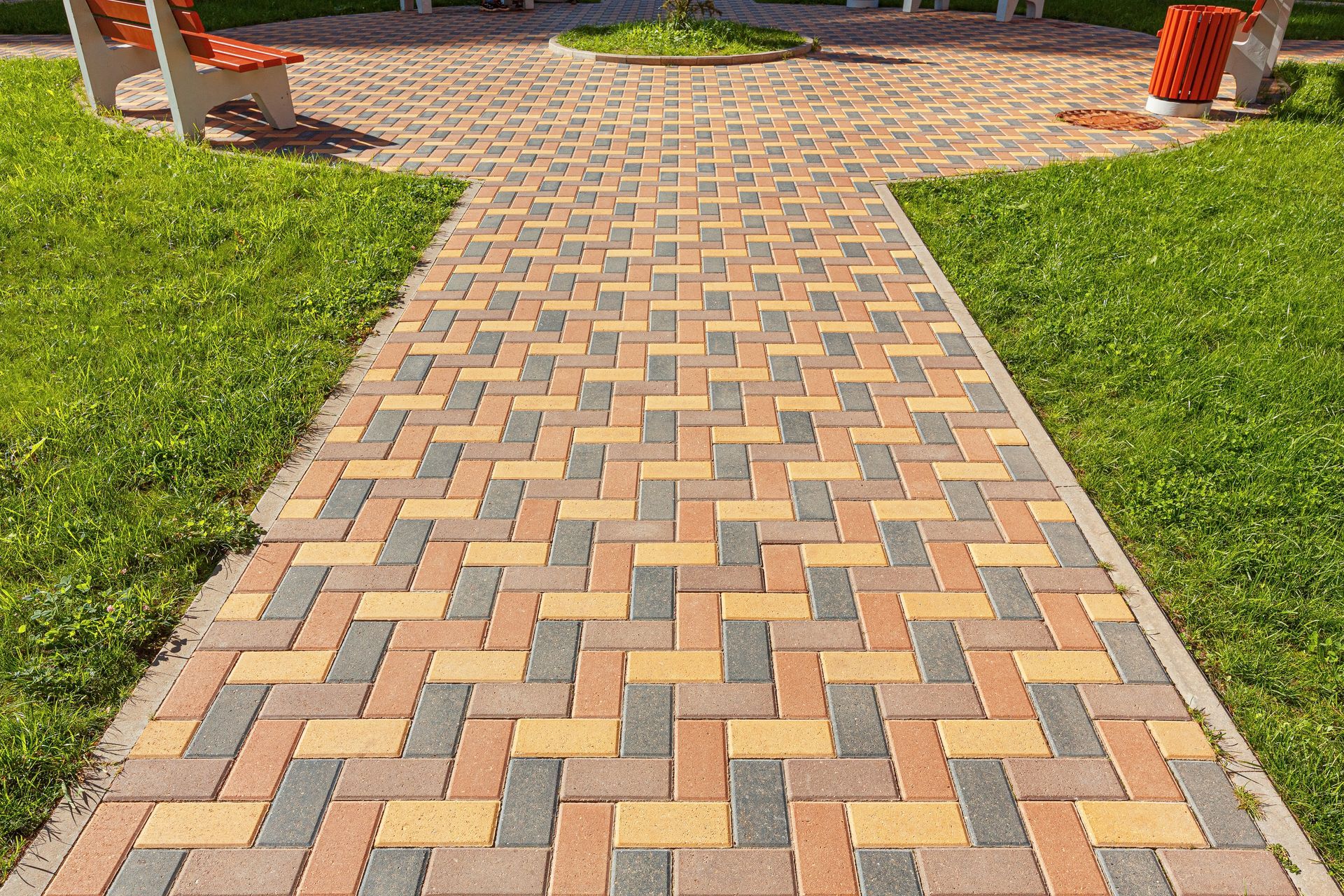 A patterned stone walkway with grey, tan, and orange rectangular pavers between two patches of grass in a park.