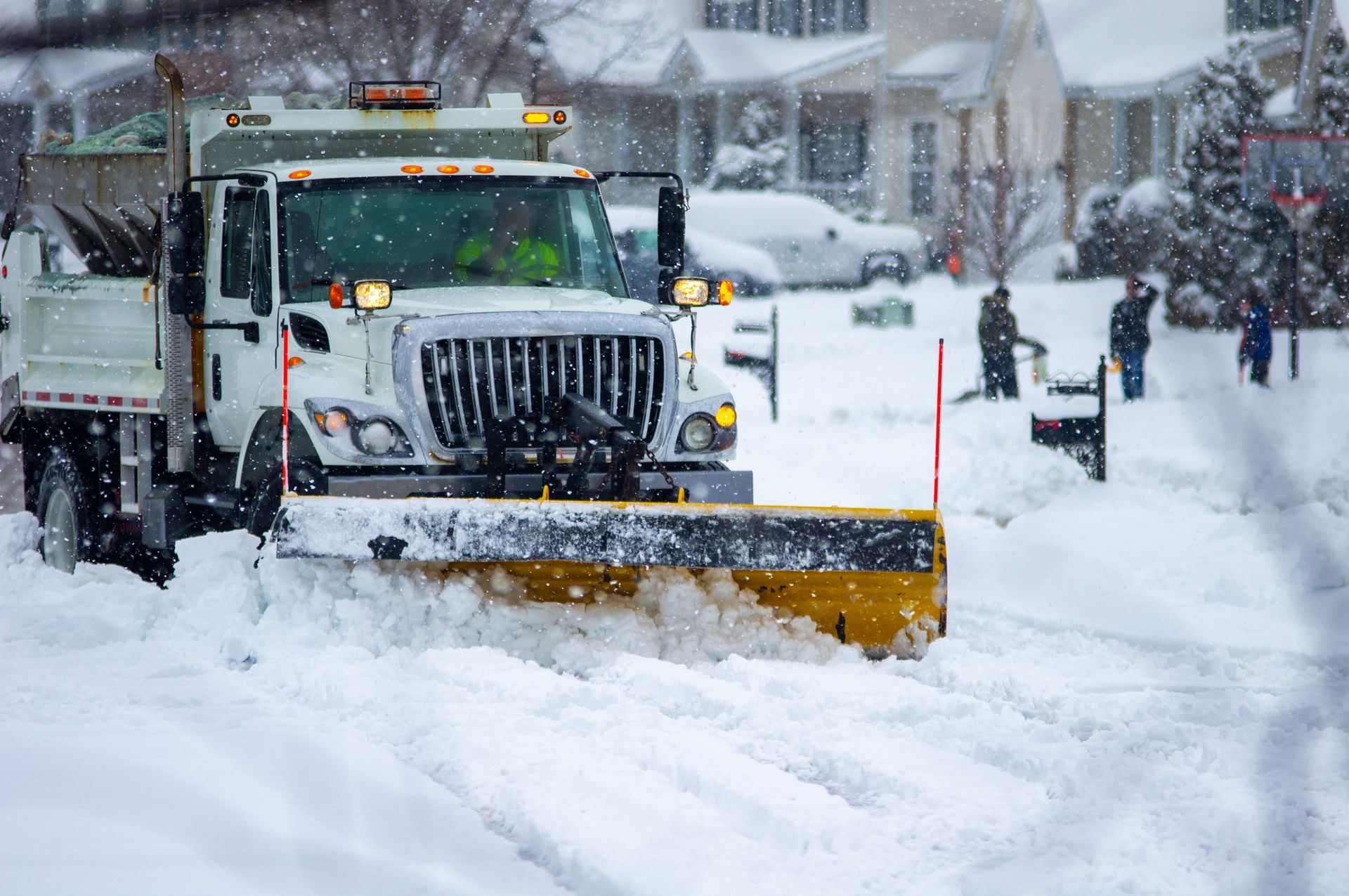A white snowplow clears a snow-covered suburban street as people clear their driveways in the background.