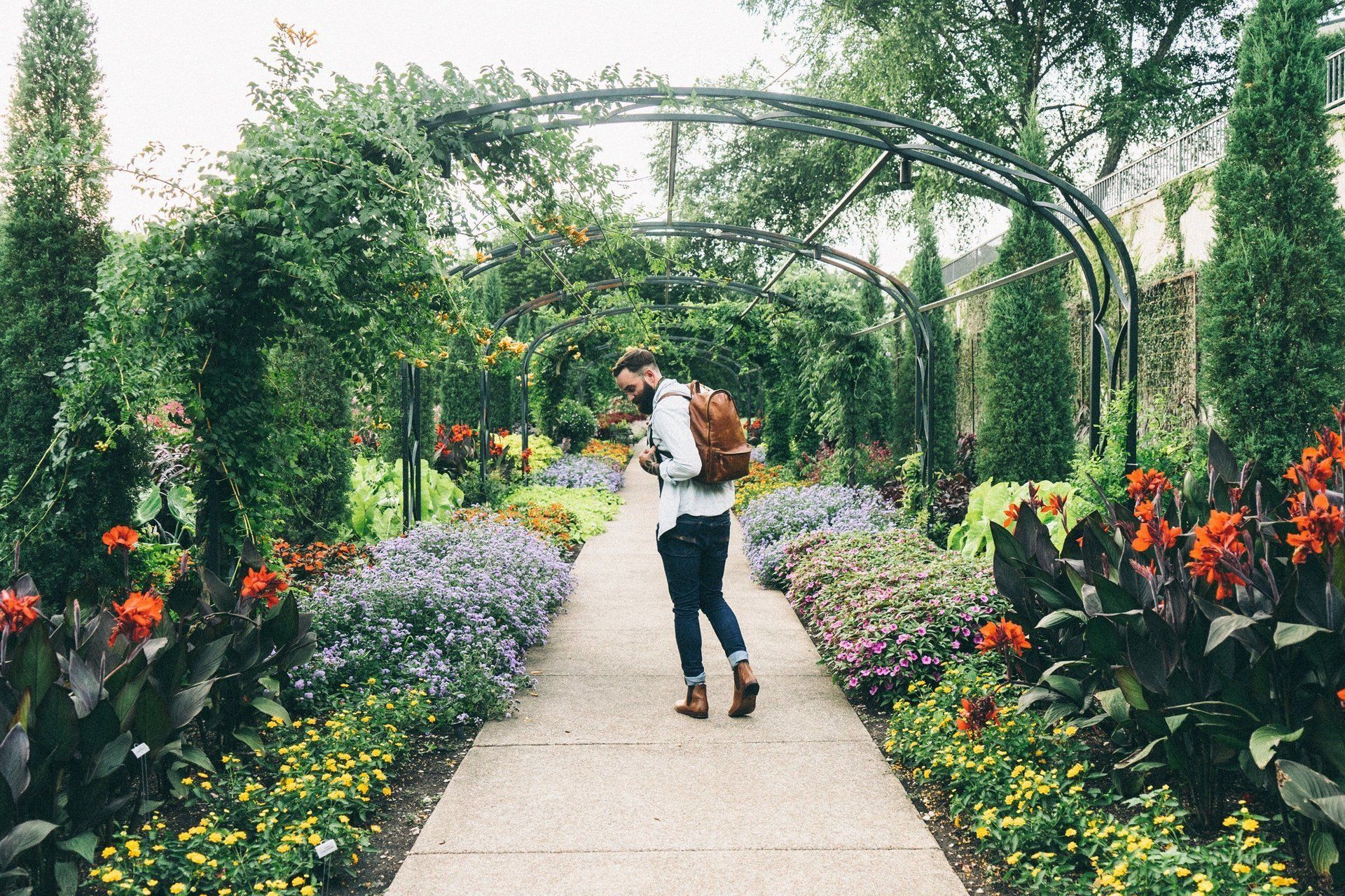 A person with a brown backpack walks down a stone path through a lush, archway-covered garden filled with colorful flowers.