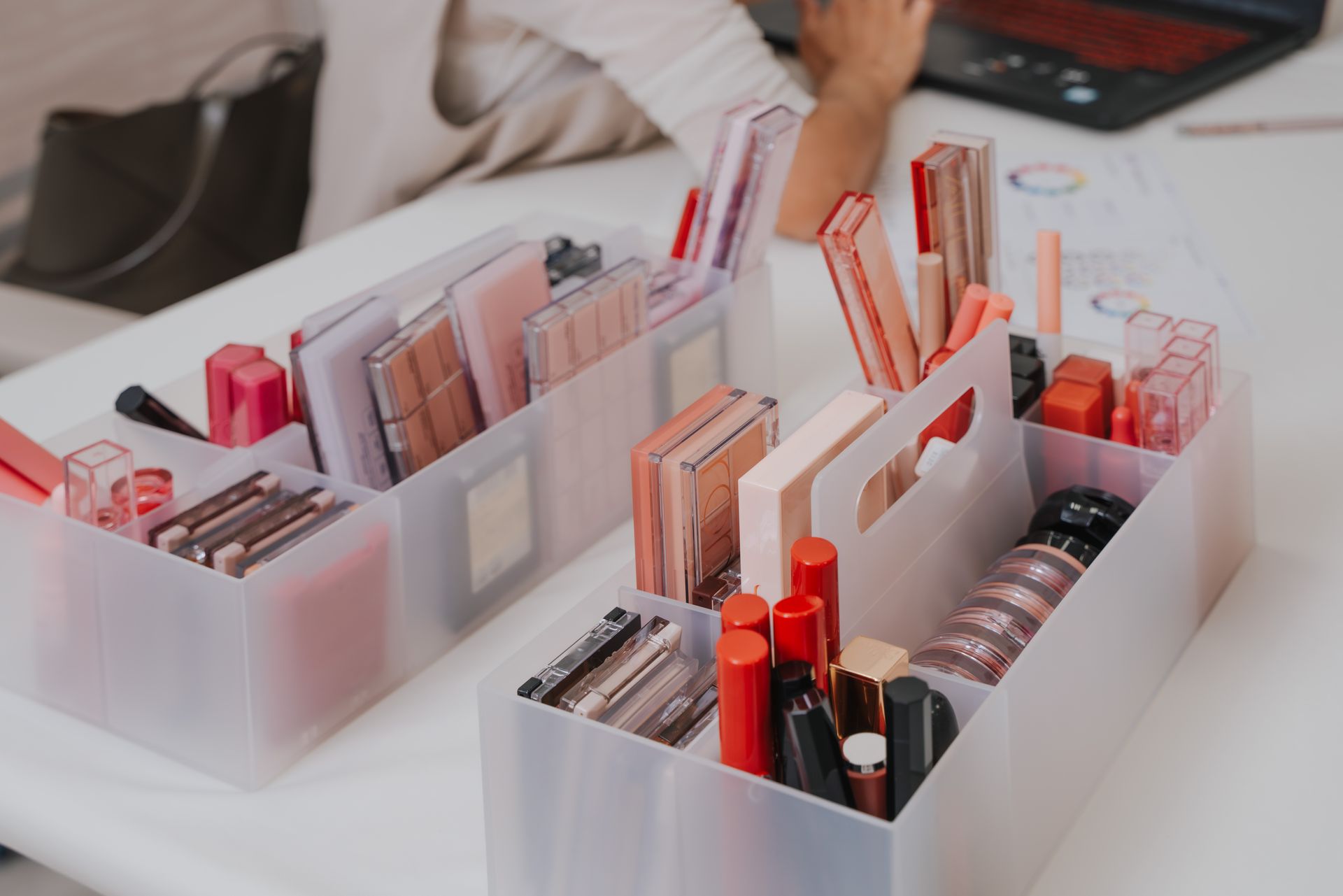 一位女士坐在辦公桌前，手裡拿著一台筆記型電腦，塑膠容器裡放著一堆化妝品。